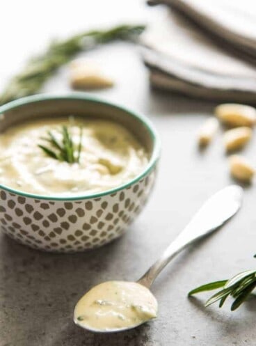 a bowl and a spoon filled with garlic and rosemary aioli with fresh garlic cloves and rosemary scattered around