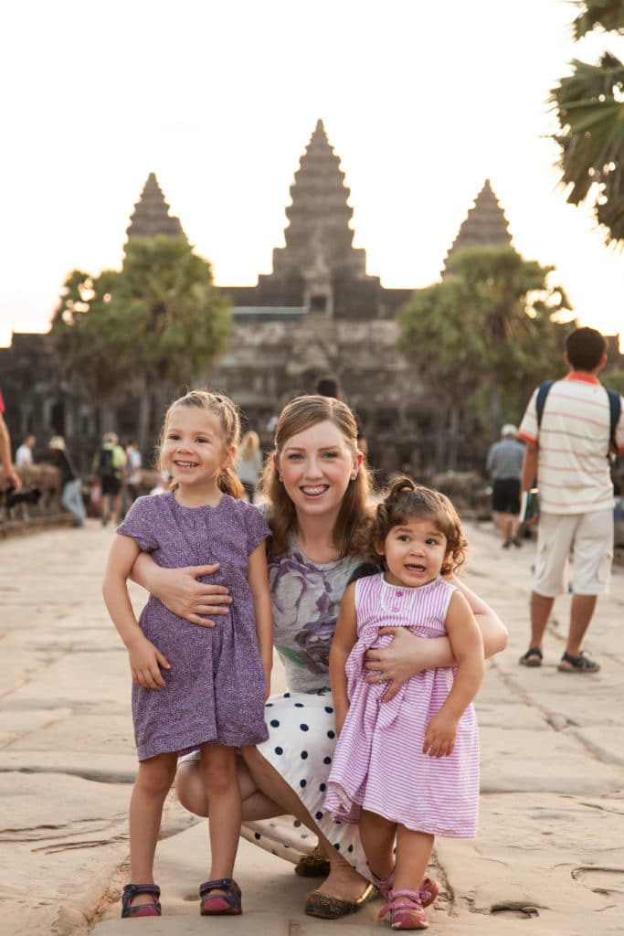 A woman and two young girls standing in front of a large Cambodian monument building with tourists in the background
