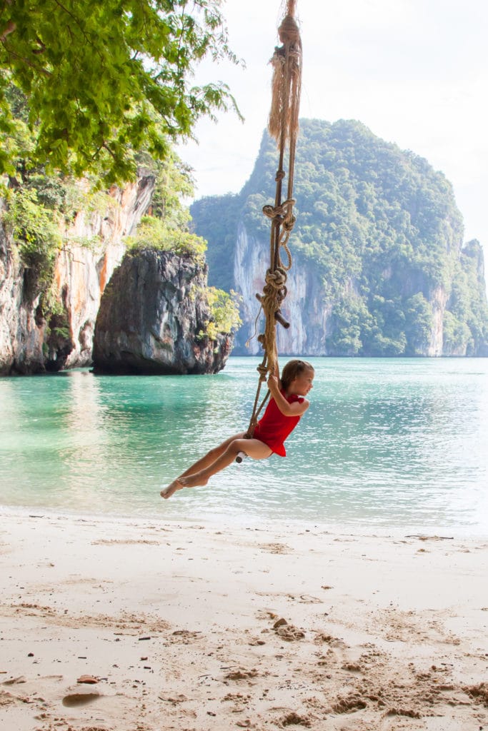 A young girl in a red dress swinging on a rope swing on the beach looking out into the water