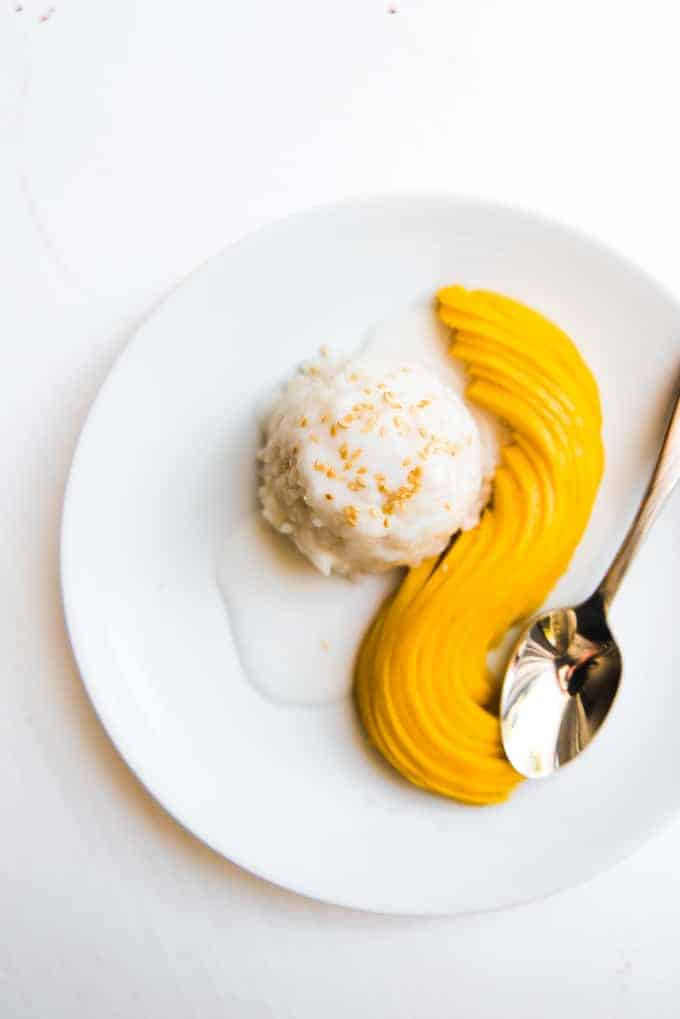an aerial view of a scoop of sticky rice covered in sauce next to some neatly arranged mango slices and a shiny spoon