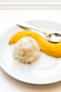 a white plate with a round scoop of sticky rice covered in sauce next to slices of mango neatly arranges and a spoon to the side
