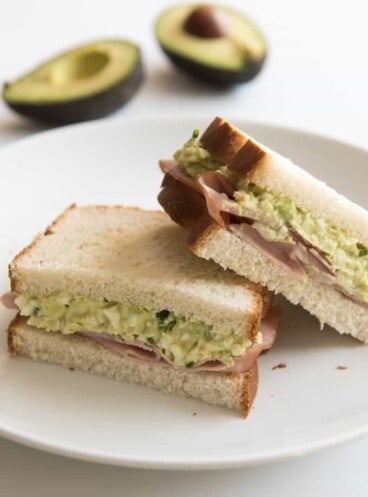 two cut halves of a green eggs and ham sandwich on a plate with halved avocados in the background