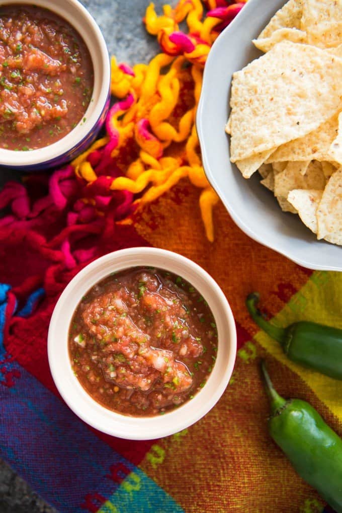 bowls of salsa next to peppers and a bowl of tortilla chips on a colorful cloth