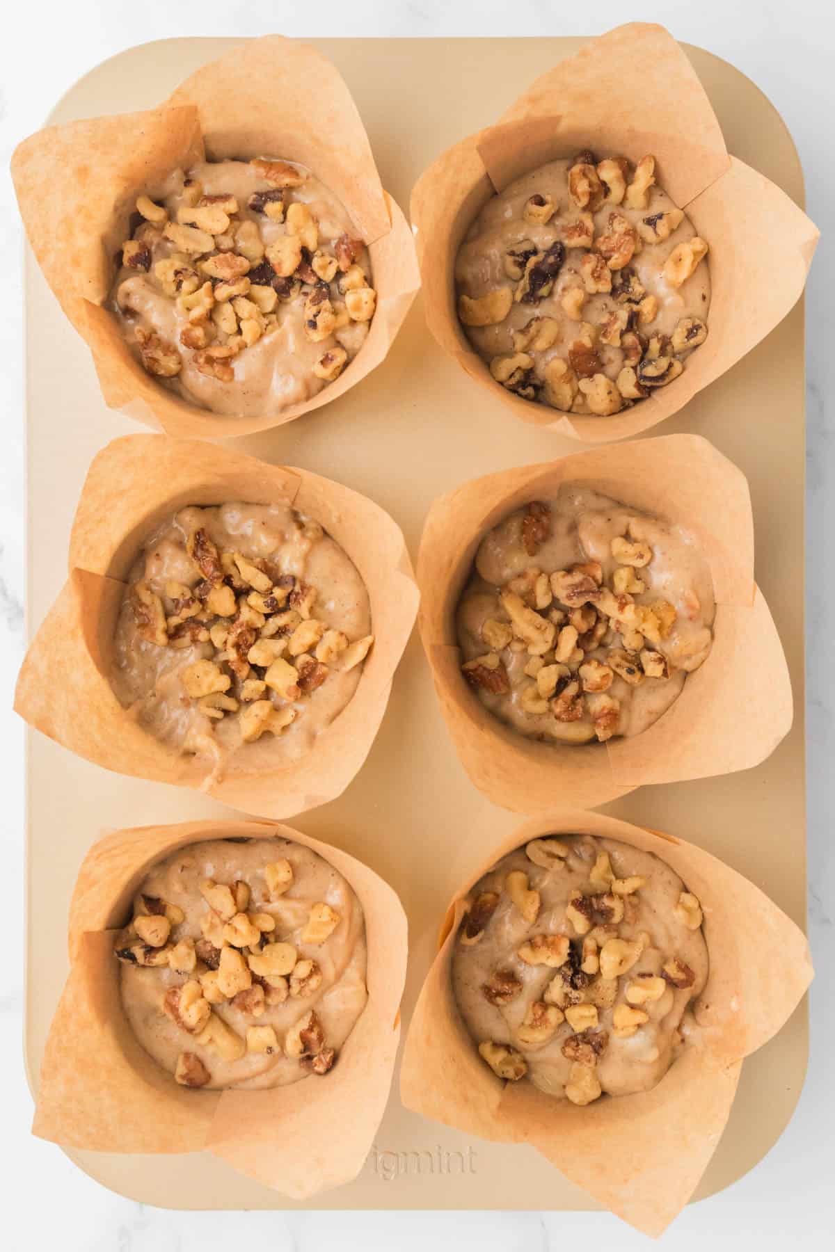 An overhead image of banana nut muffin batter in a muffin pan with paper liners and chopped walnuts sprinkled on top.