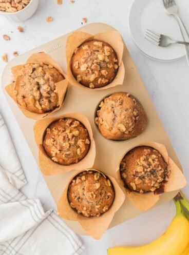 An overhead image of banana nut muffins on a pan next to a plate with forks, a cloth napkin, and bananas.