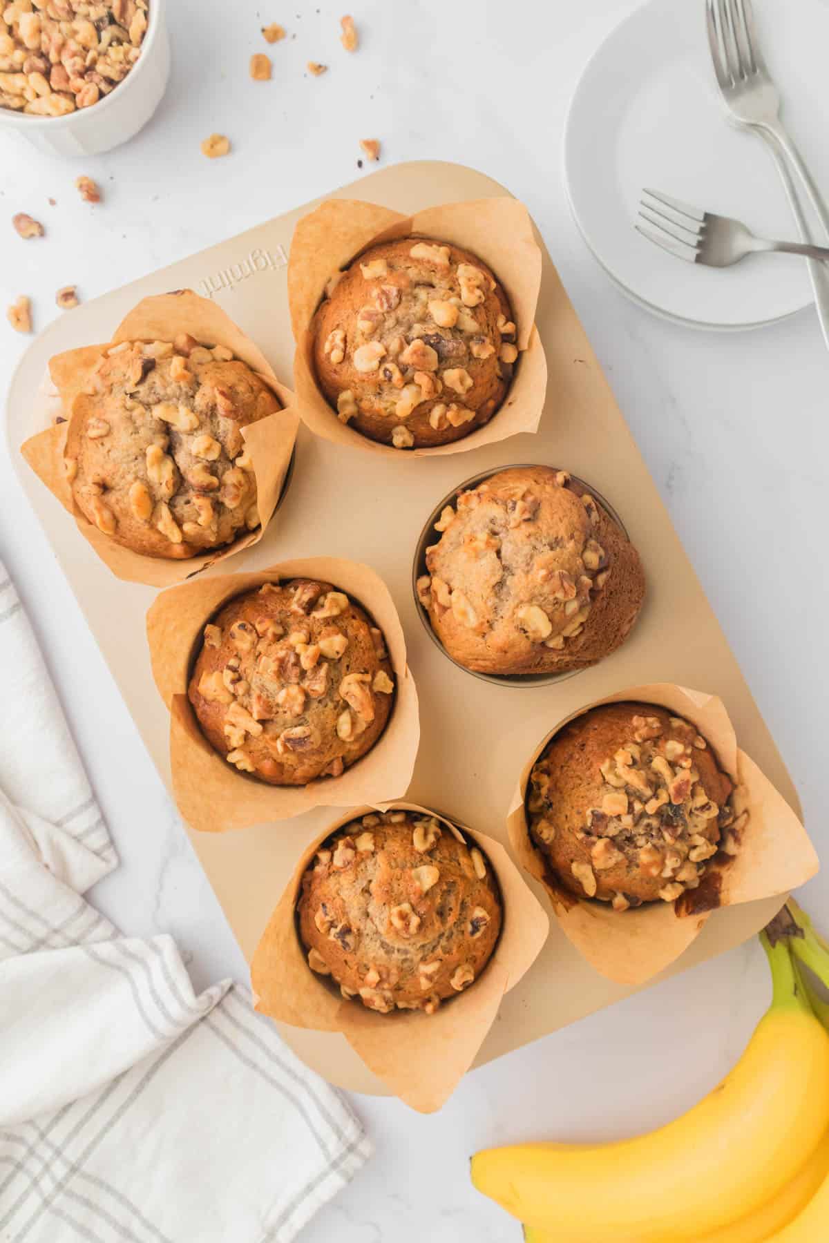 An overhead image of banana nut muffins on a pan next to a plate with forks, a cloth napkin, and bananas.