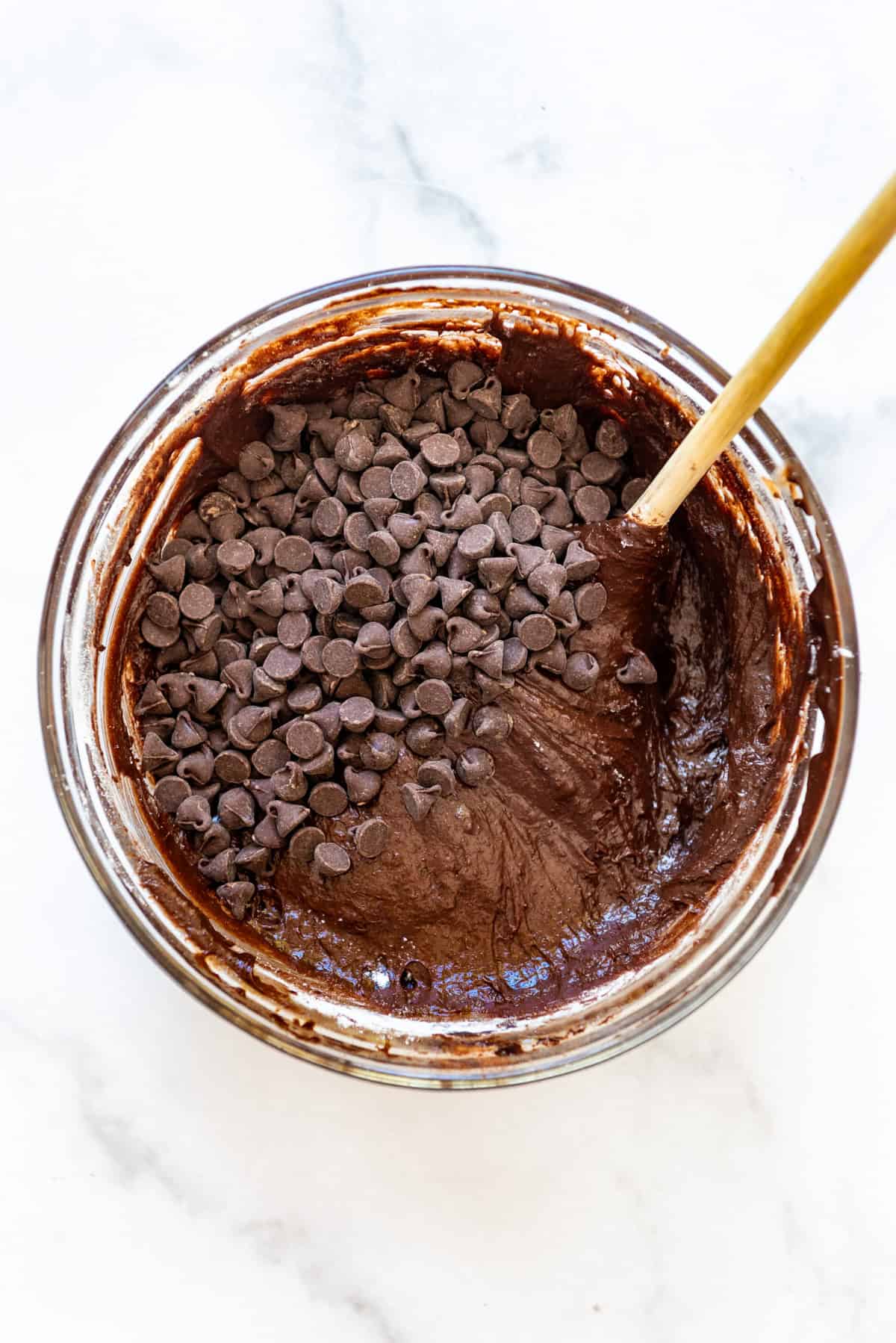 Brownie batter in a glass bowl with chocolate chips being stirred in with a wooden spoon.