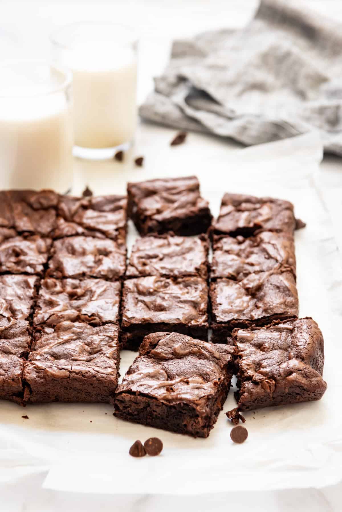 Brownie squares on parchment paper next to glasses of milk.