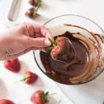 a hand holding a chocolate dipped strawberry over a bowl of melted chocolate