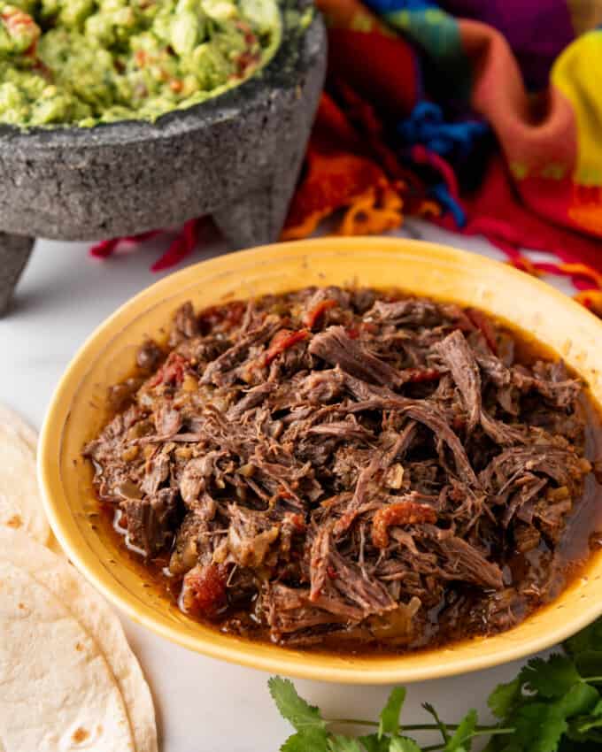 An image of a yellow bowl full of shredded beef with Mexican spices in front of a bowl of guacamole.