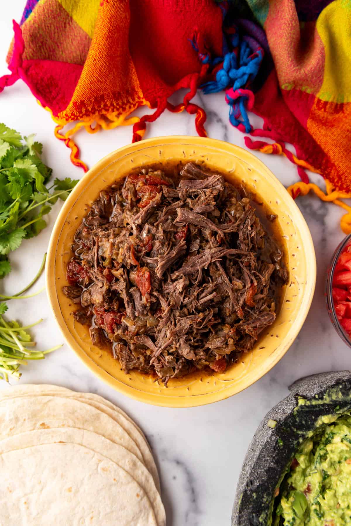 An image of a bowl of Mexican shredded beef surrounded by tortillas, cilantro, and guacamole.