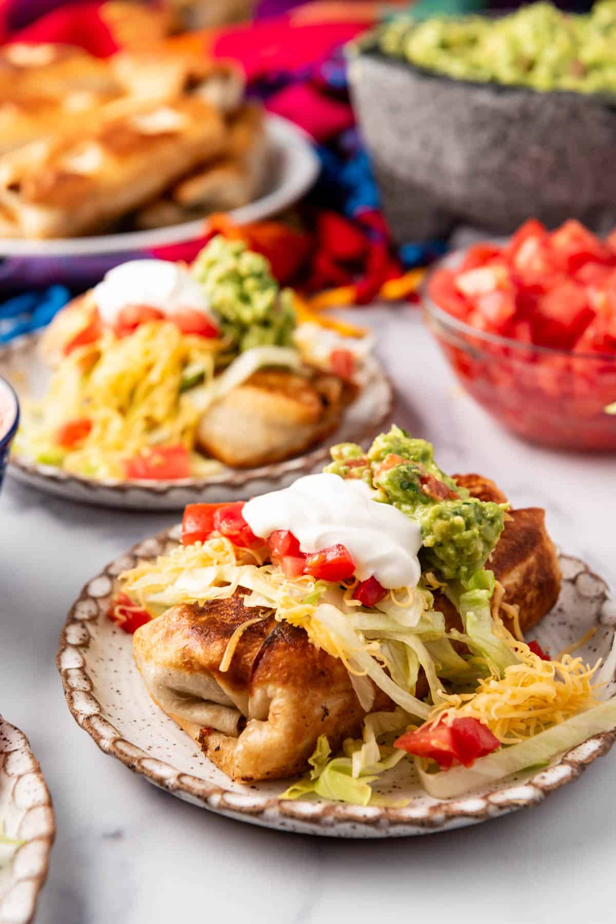 An image of shredded beef chimichangas on plates topped with sour cream, guacamole, cheese, shredded lettuce, and tomatoes.