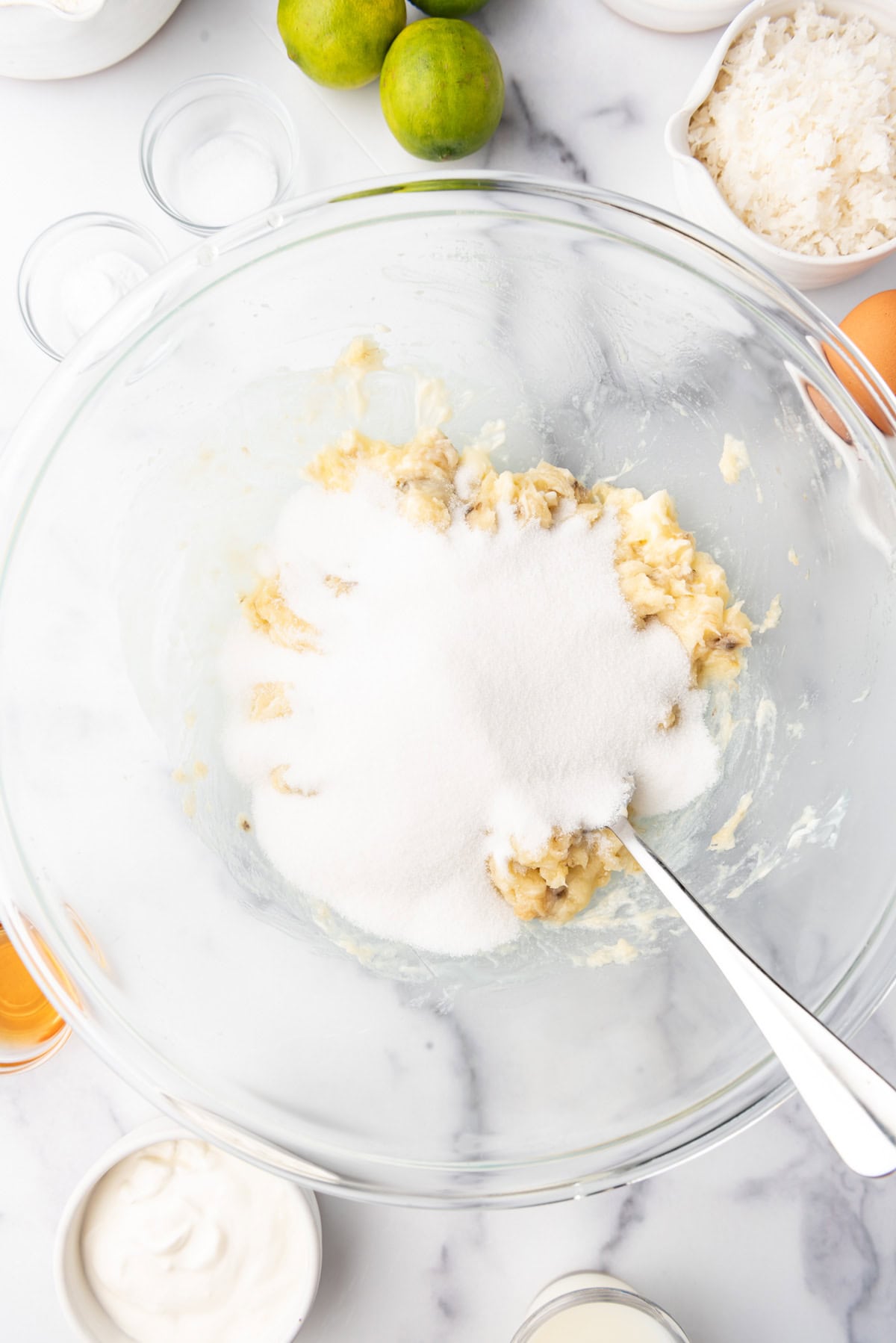 Adding sugar to mashed bananas and butter in a glass mixing bowl.