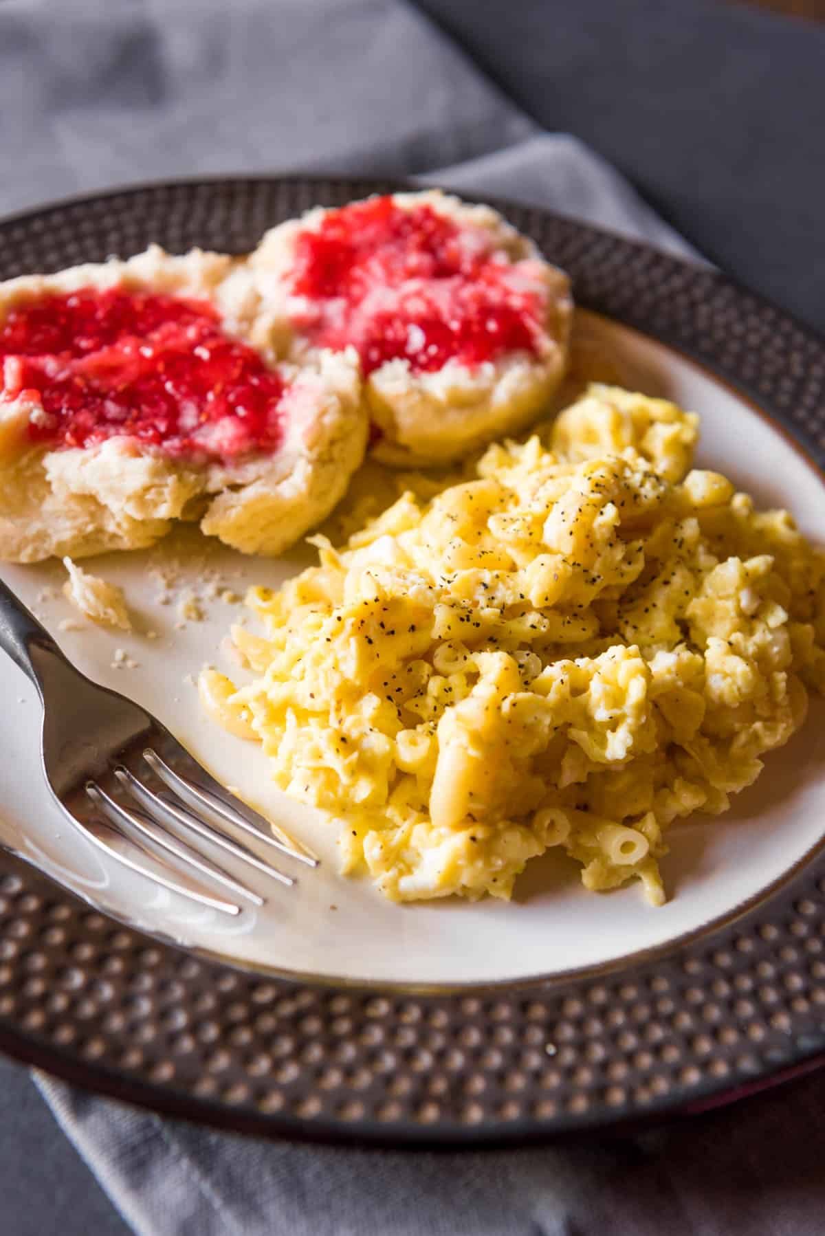 Peppery scrambled macaroni eggs on a plate with a fork and jam covered biscuit.