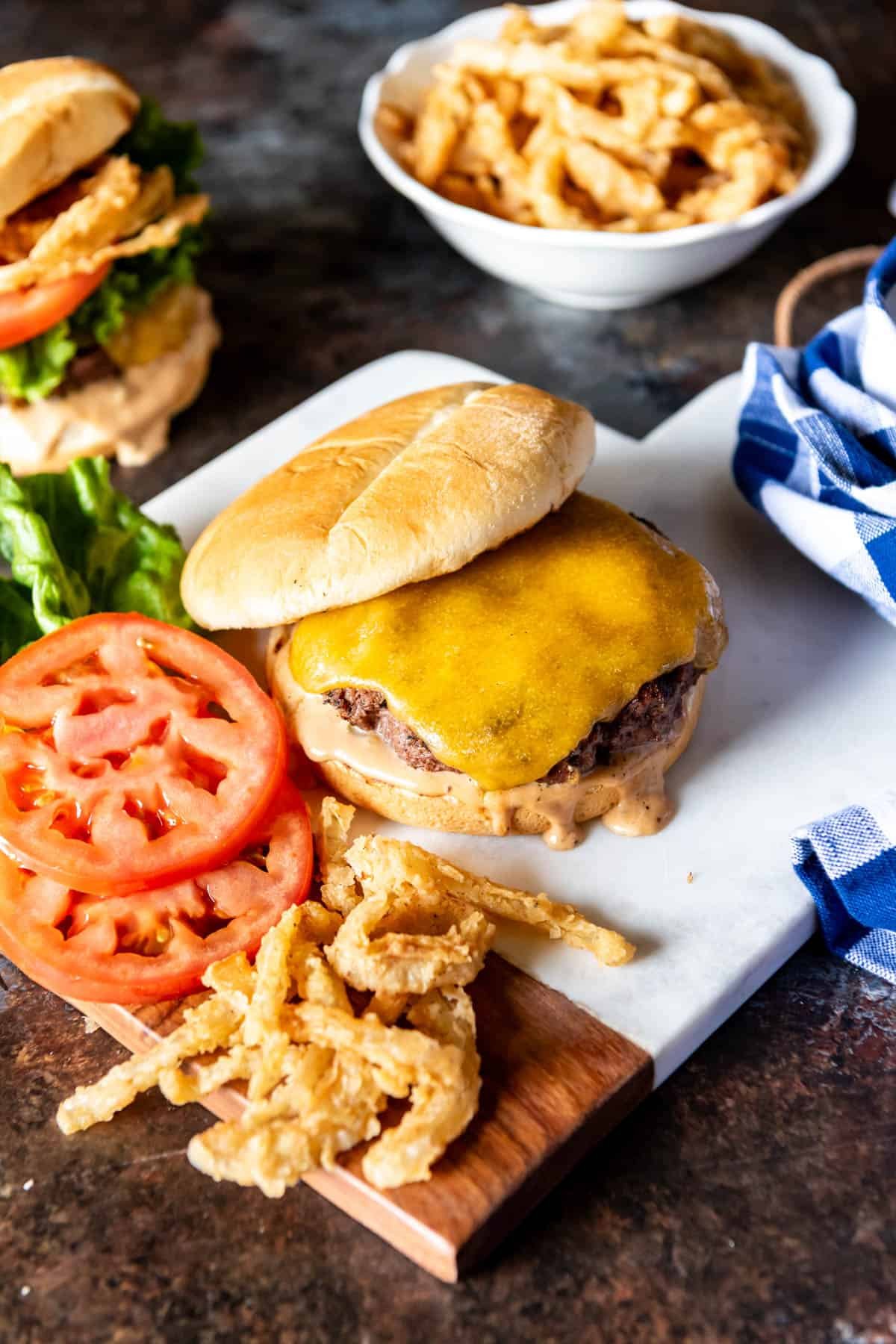A cheeseburger with the top bun slid over to show melted cheese and crispy onion strings sliced tomatoes and lettuce off to the side.
