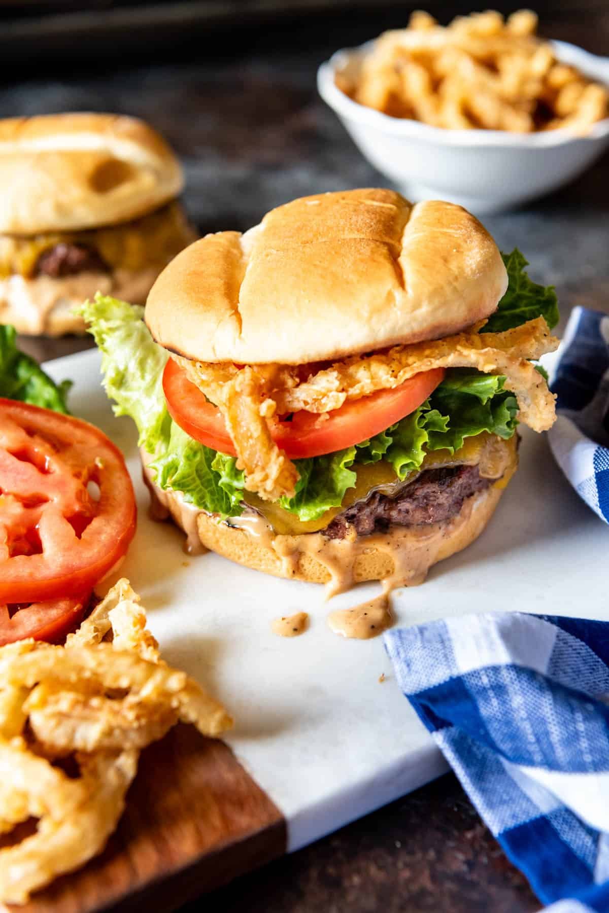A burger with crispy onion strings and sauce dripping onto the board below.