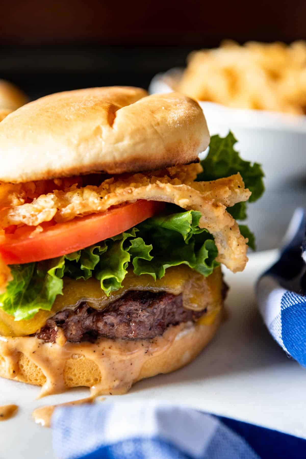 An all american burger with crispy onion strings tomato lettuce cheese and sauce dripping.