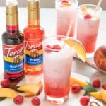 two bottles of torani syrup next to 3 glasses full of freshly made raspberry peach italian soda with fresh raspberries and peached scattered all around and garnishing the cups