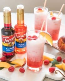two bottles of torani syrup next to 3 glasses full of freshly made raspberry peach italian soda with fresh raspberries and peached scattered all around and garnishing the cups
