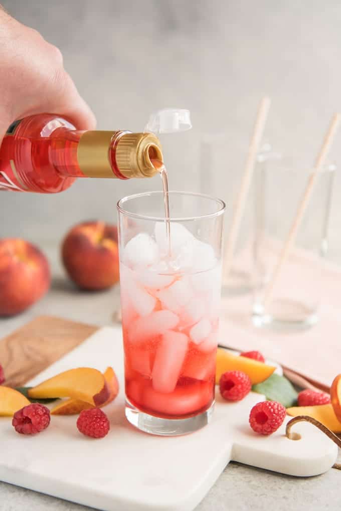 pouring syrup into a glass of ice surrounded by fresh peaches and raspberries