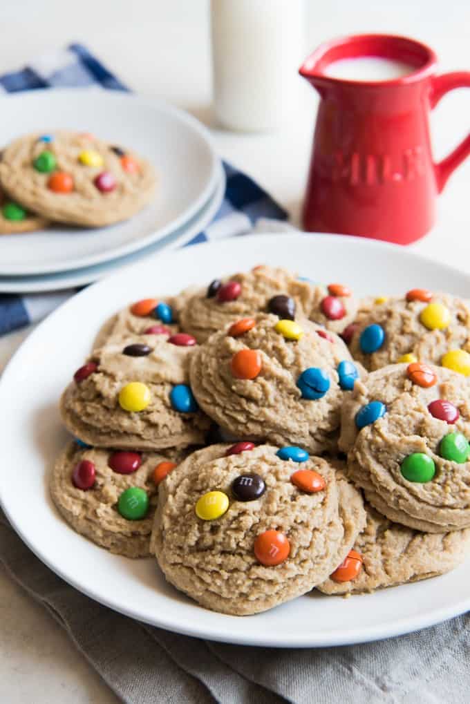 big and soft m&m cookies on ahwite plates with a pitcher of milk to the side
