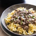 A gray plate contains cooked egg noodles topped with freshly made beef strongaff with mushrooms and a skillet in the background holds more sauce