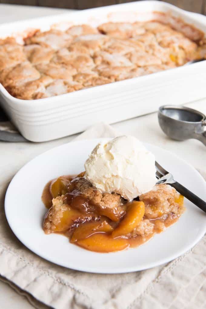 a baking dish behind a white plate of peach cobbler and a scoop of ice cream