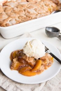 peach cobbler in a baking dish and a servings worth on a white plate in front and topped with ice cream