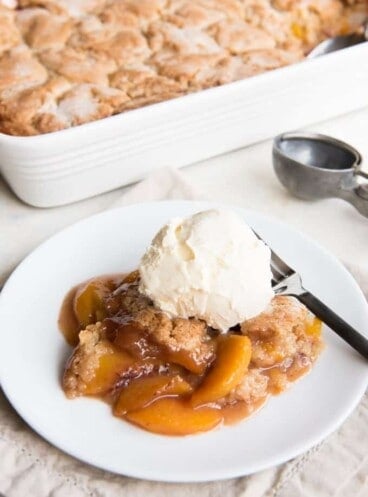 peach cobbler in a baking dish and a servings worth on a white plate in front and topped with ice cream