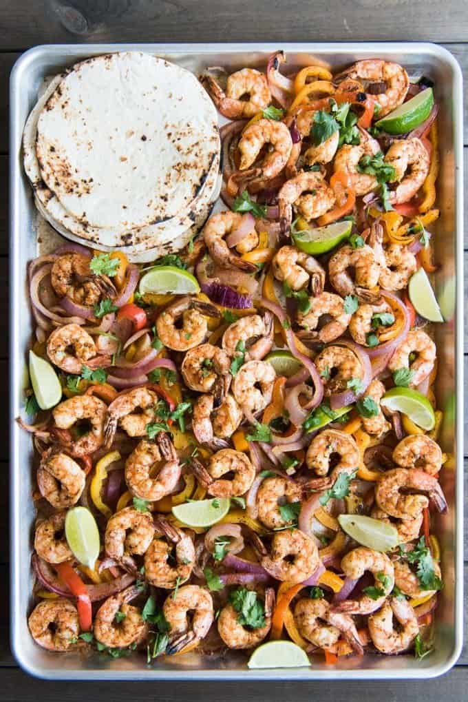 shrimp fajita filling and lime wedges on a baking sheet with a stack of tasted tortillas
