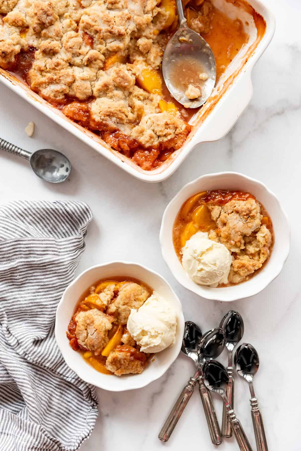 An overhead image of two bowls of peach cobbler next to a baking dish and spoons.