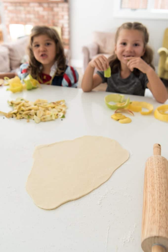 kinderen aan een toonbank naast stapels appelschillen met een uitgerold deeg ervoor