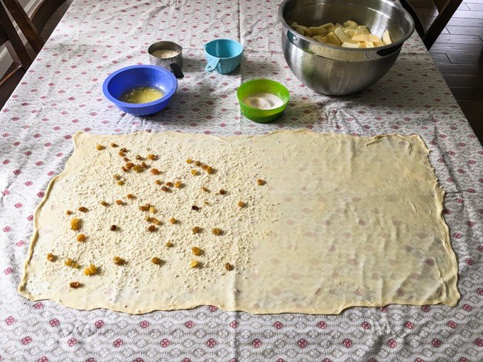 Zeer dunne rechthoek van deeg uitgerold op een tafelkleed met andere ingrediënten in kommen erachter. sommige ingrediënten zijn op de helft van het deeg gestrooid