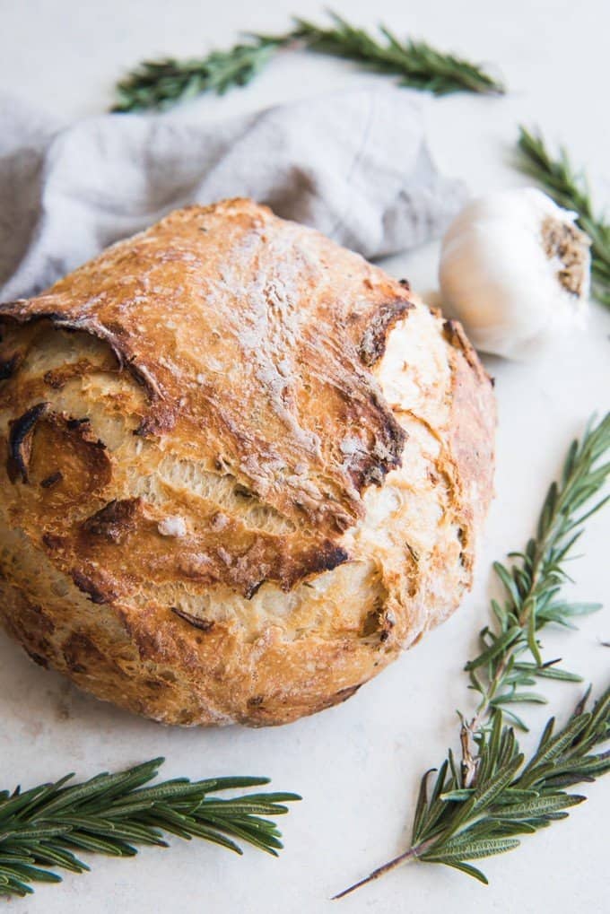 a round loaf of garlic rosemary artisan bread surrounded by fresh rosemary and garlic head