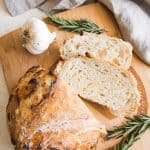 Roasted Garlic & Rosemary No Knead Artisan Bread on a wooden cutting board with fresh rosemary and garlic