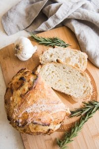 Roasted Garlic & Rosemary No Knead Artisan Bread on a wooden cutting board with fresh rosemary and garlic