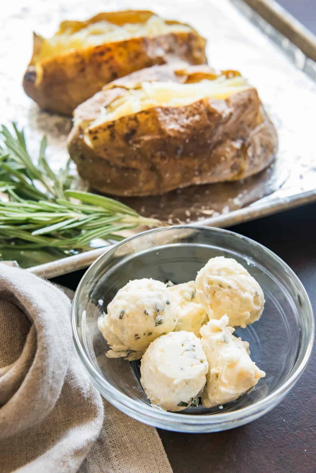 Balls of roasted garlic & rosemary compound butter in a bowl with baked potatoes sliced open on a baking sheet in the background.