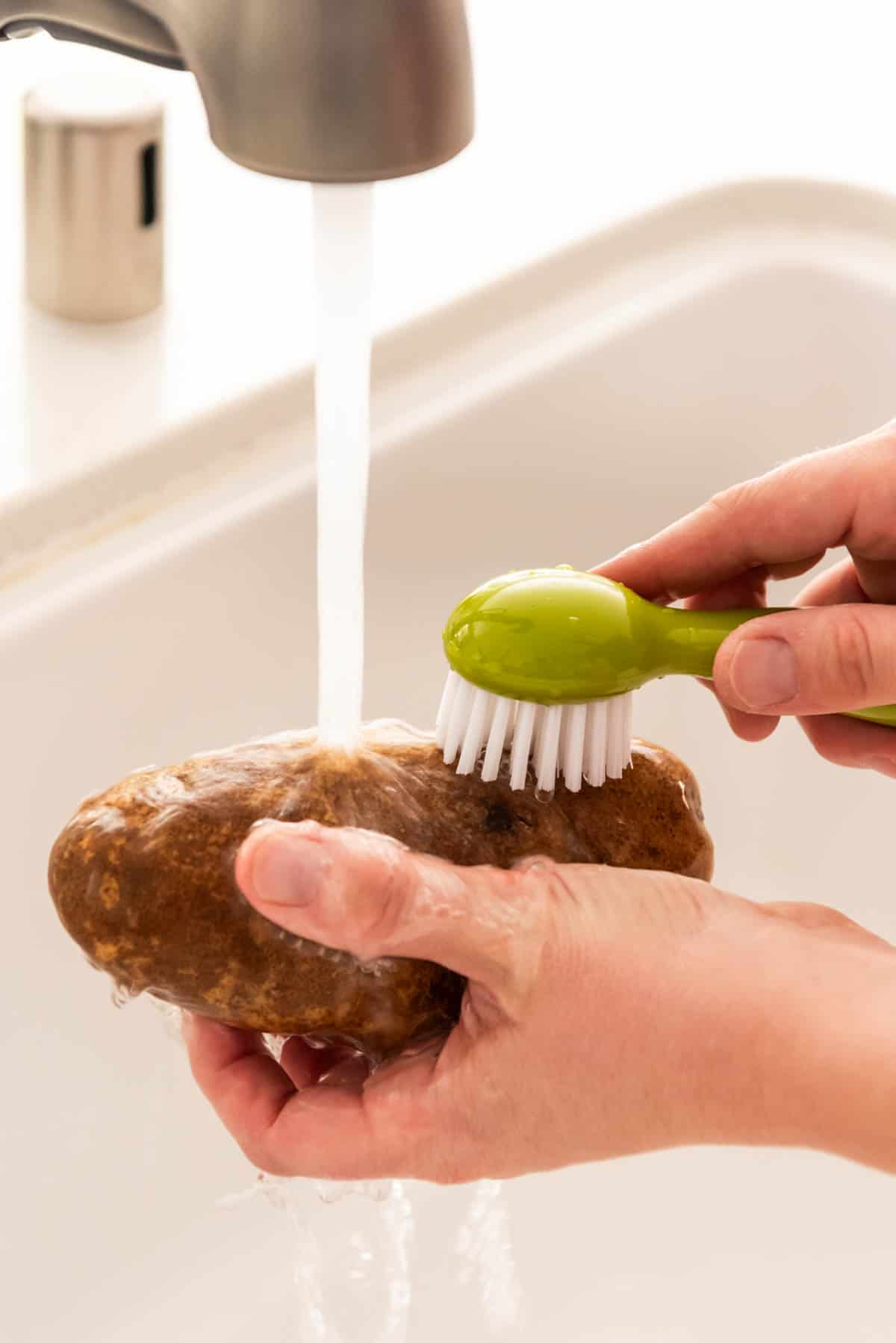 Hand holding a russet potato under running water with a scrub brush to clean off any dirt before baking.