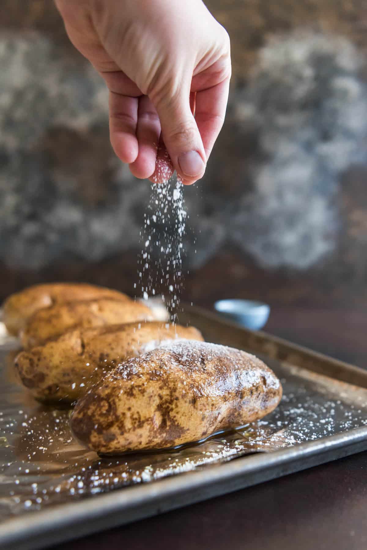 Hand drizzling coarse salt over an oil rubbed russet potato.