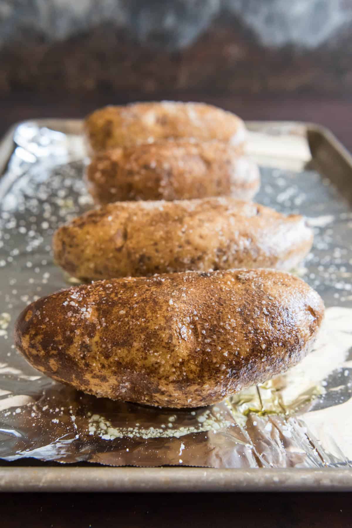 Four large russet potatoes on a foil lined baking sheet, rubbed with oil and salt and ready to go into the oven.