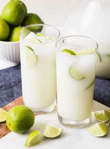two glass cups full of brazillian limade and ice with limes and lime wedges around them