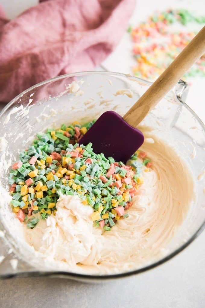 Mixing bowl with cream cheese frosting and rainbow chips poured in ready to be mixed together with a spatula.