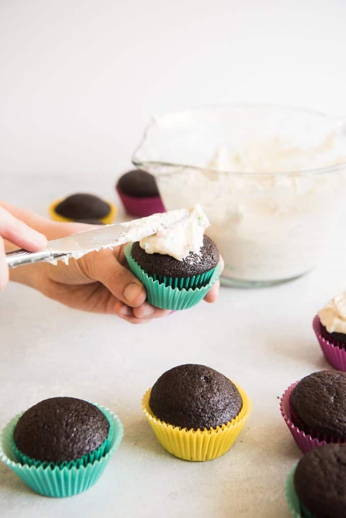 Hands holding a knife being used to frost chocolate cupcakes with homemade rainbow chip frosting.