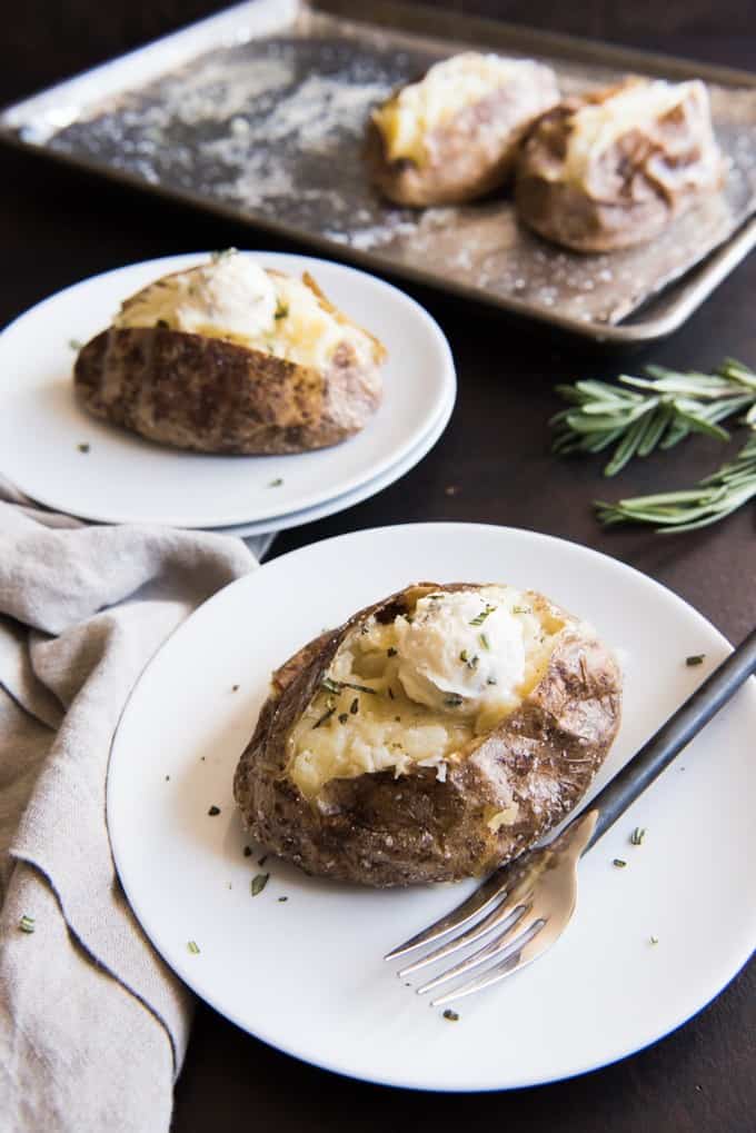 Salt Crusted Baked Russet Potatoes split open and sitting on plates with roasted garlic and rosemary compound butter on top.