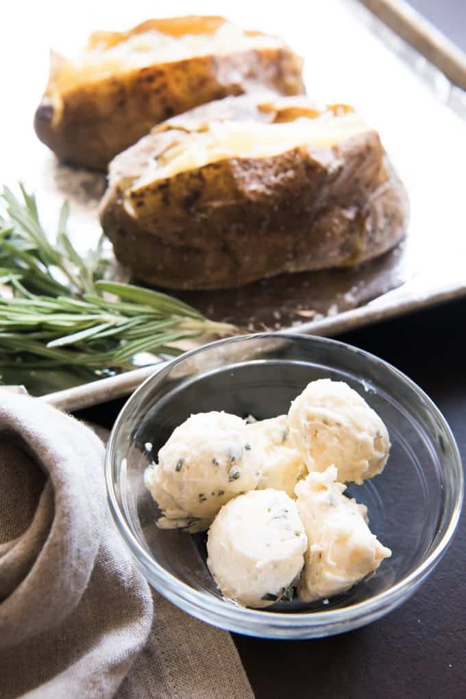 Balls of roasted garlic & rosemary compound butter in a bowl with baked potatoes sliced open on a baking sheet in the background.