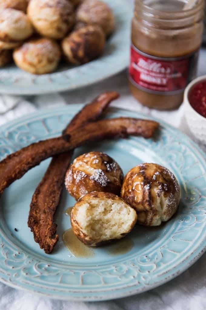 Danish aebleskiver (or ebelskiver) dusted with powdered sugar, drizzled with honey, and served on a plate with a rasher of bacon.