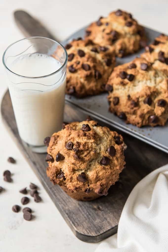 baked chocolate chip muffins in a baking dish with one out and to the side on a wooden cuttting board with a glass of milk next to some scattered chocolate chips