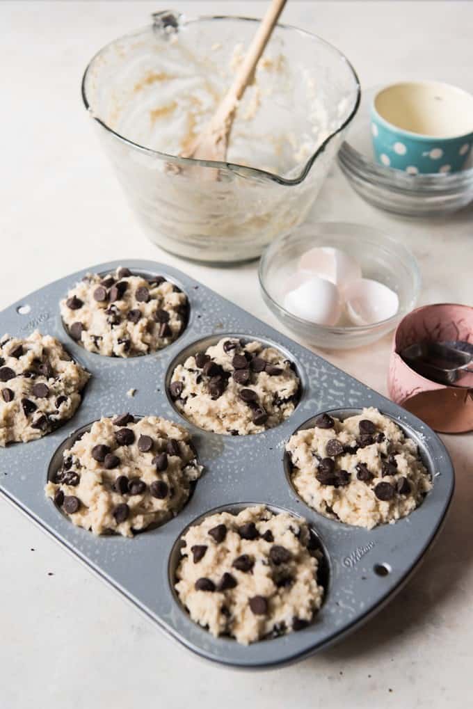 A muffin pan with raw chocolate chip muffin batter in each cup ready to be baked and the used tools and egg shells off to the side
