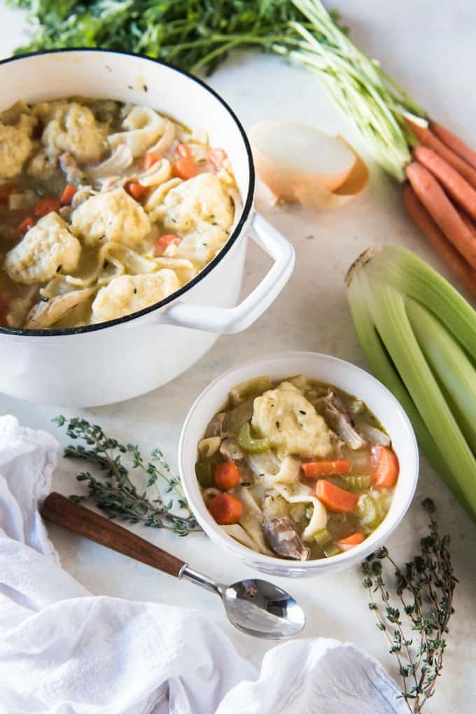 A bowl of chunky chicken noodle soup with dumplings and a pot of soup behind it with carrots, onions, and celery surrounding them.
