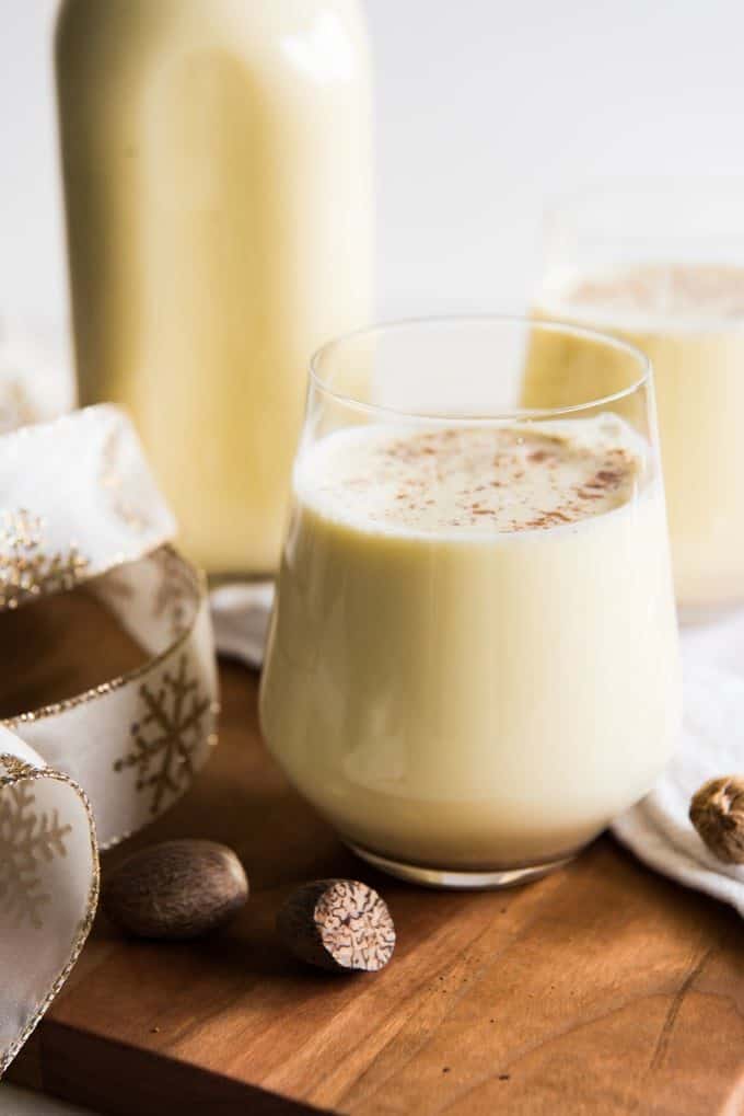 a glass of fresh homemade eggnog in front of a another glass full and a jug of eggnog in back. Ribbons and nuts rest around it.